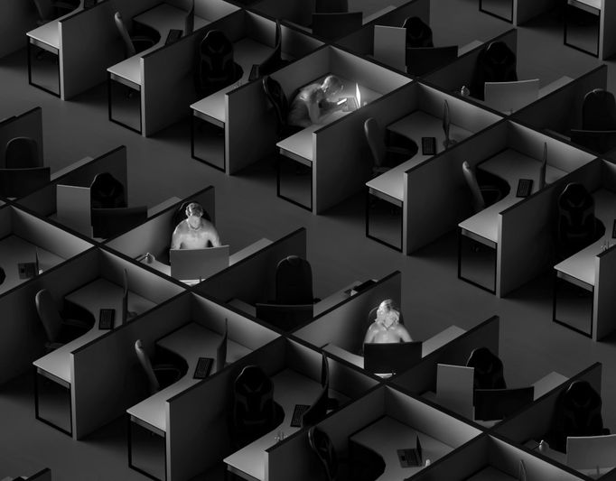 Image monochrome d'un bureau faiblement éclairé avec des personnes travaillant dans des cabines isolées, chacune éclairée par leurs écrans d'ordinateur.