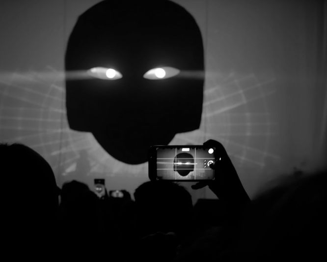 Silhouette of a large face with glowing eyes projected on a screen, while a person records it on a smartphone in a dark room.