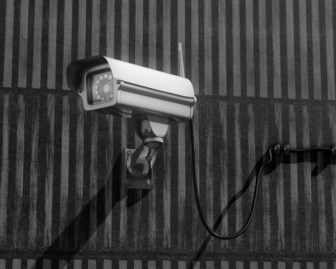 Black and white image of a security camera mounted on a striped wall, casting a shadow.