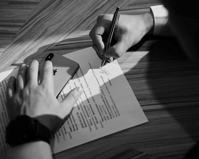 Una persona firma un documento en una mesa de madera, sosteniendo un bolígrafo con una mano y un teléfono inteligente con la otra.