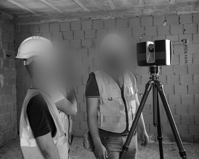 Dos trabajadores de la construcción con chalecos de seguridad y un casco operan un instrumento de topografía montado en un trípode dentro de un edificio de ladrillo en construcción.