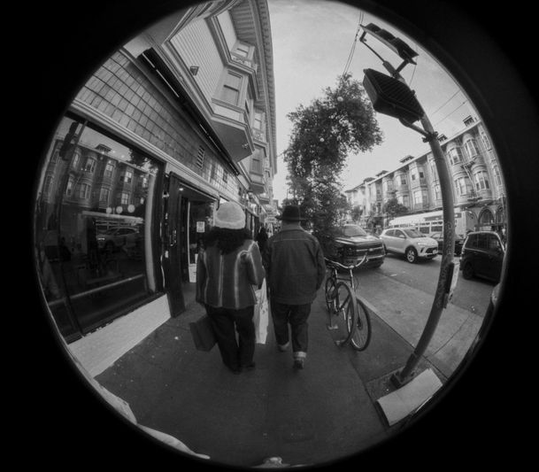 Fish-eye view of a city street with two people walking on the sidewalk, buildings, parked cars, and a bicycle in the background.