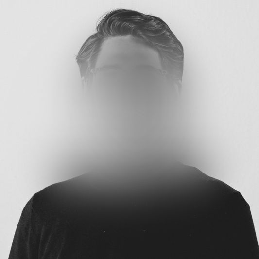 Une personne aux cheveux courts et foncés, vêtue d'une chemise noire. Son visage est masqué par un effet gris, semblable à du brouillard.