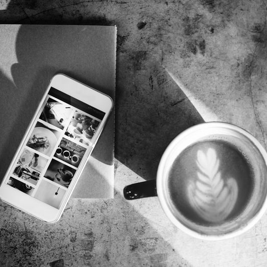 A smartphone displaying photos rests on a notebook, next to a cup of coffee with latte art, casting a shadow on a textured surface. Black-and-white image.