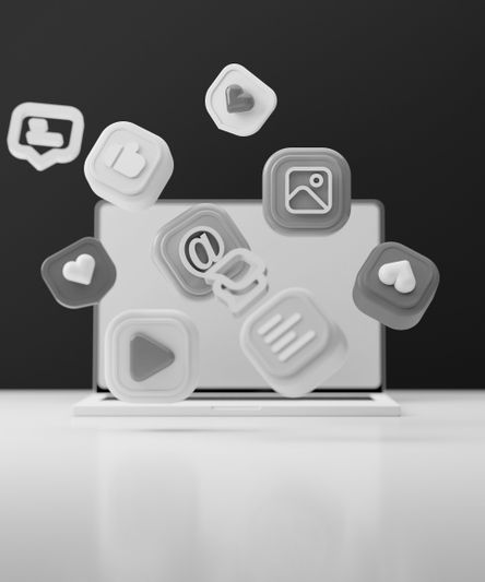 Icônes de médias sociaux 3D flottantes en niveaux de gris, notamment un cœur, un symbole @ et un bouton de lecture, devant un ordinateur portable ouvert sur une surface blanche.