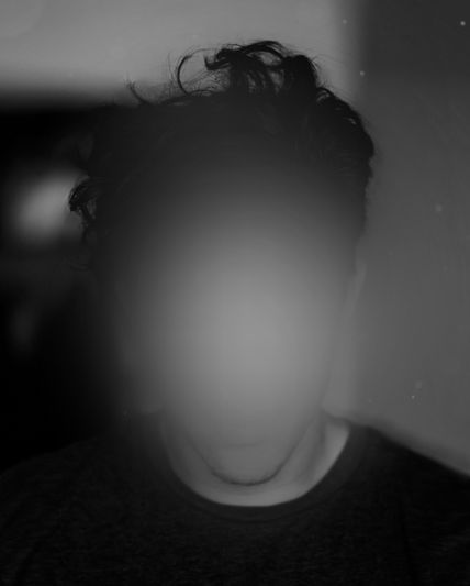 Black-and-white portrait of a person with tousled hair and their face obscured by a soft blur, wearing a dark shirt against a dim background.