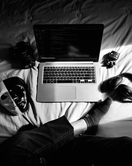 Image monochrome d'un ordinateur portable avec du code à l'écran, entouré d'une caméra, de masques, d'un casque et d'une figurine. On distingue le pied d'une personne.