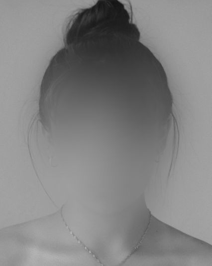 Rostro borroso de una persona con un moño, que lleva pequeños pendientes de aro y un delicado collar, sobre un fondo liso.