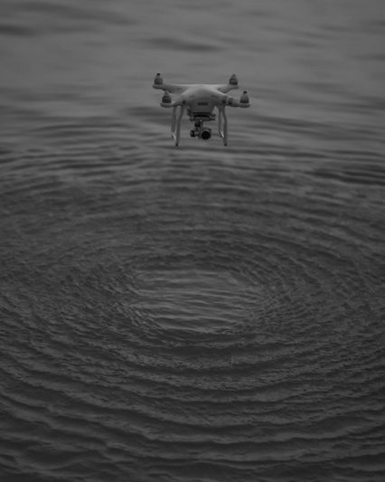 A drone hovers above rippling water, creating circular patterns on the surface. The image is in grayscale.