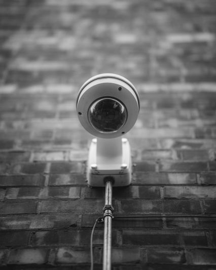 Black and white image of a security camera mounted on a brick wall, viewed from below, with focus on the camera's round lens.