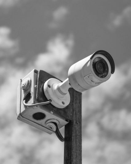 Black and white image of a security camera mounted on a pole against a cloudy sky background.