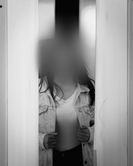 Une personne au visage flou se tient entre deux grandes structures verticales, vêtue d'une veste en jean et d'une chemise blanche. Image en noir et blanc.