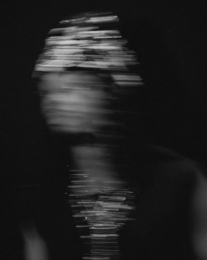 Imagen borrosa en blanco y negro de una persona con un tocado decorado, creando una sensación de movimiento y misterio contra un fondo oscuro.