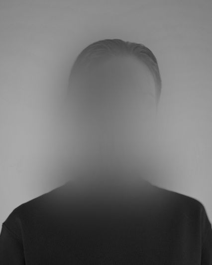 Grayscale head-and-shoulders portrait of a person in a dark shirt whose face is blurred into a soft, misty haze.
