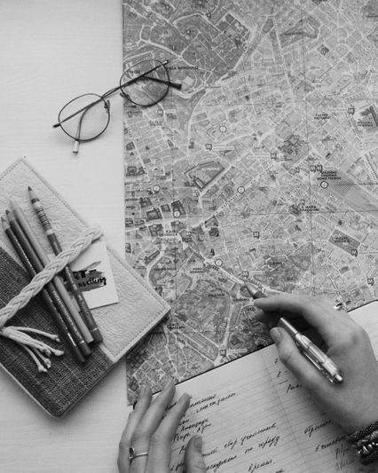 Czarno-biały obraz przedstawiający osobę studiującą szczegółową mapę miasta przy użyciu notatnika, ołówków i okularów na stole.