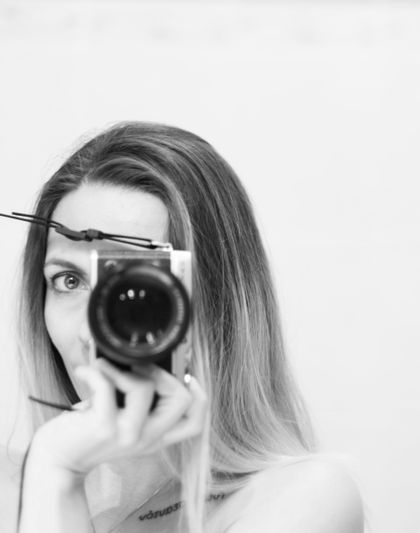 Photo en noir et blanc d'une femme tenant un appareil photo devant son visage, avec un pinceau de maquillage dans ses cheveux, capturant son reflet.