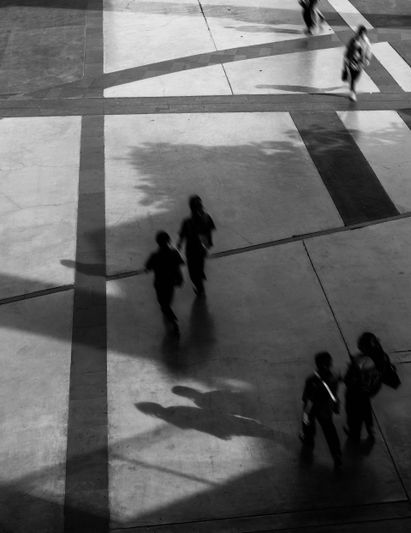 Vue aérienne de personnes marchant sur un trottoir à motifs, projetant de longues ombres au soleil. Noir et blanc.