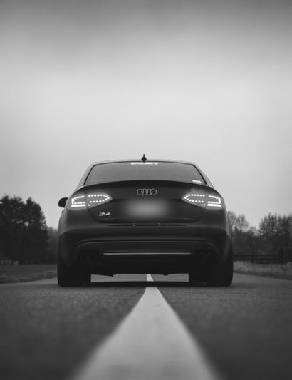 Black and white image of an Audi S4 from behind, parked on a road with a single dividing line, surrounded by trees in the distance.