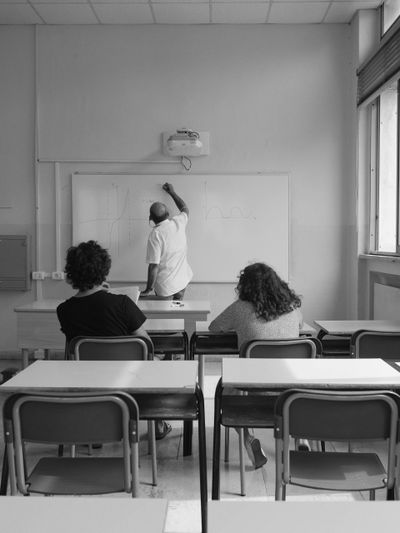 Un enseignant dessine des graphiques sur un tableau blanc dans une salle de classe, avec deux élèves assis et observant, en noir et blanc.