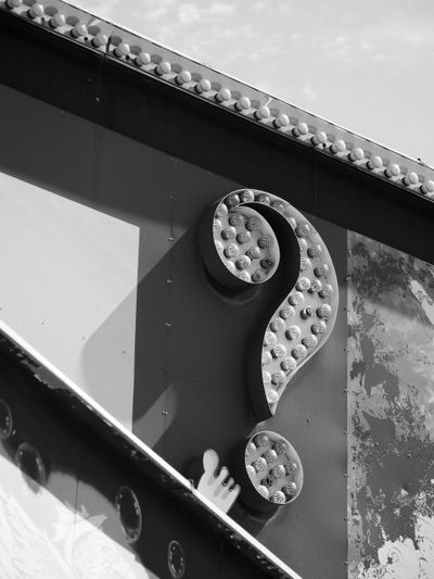 Imagen en blanco y negro de un gran cartel de signo de interrogación de estilo vintage con bombillas, colocado contra una pared parcialmente sombreada.