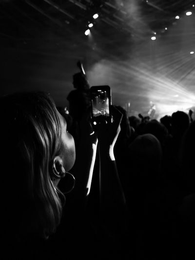 Person in a crowd capturing a concert on their phone, with dramatic stage lighting and beams illuminating the scene.