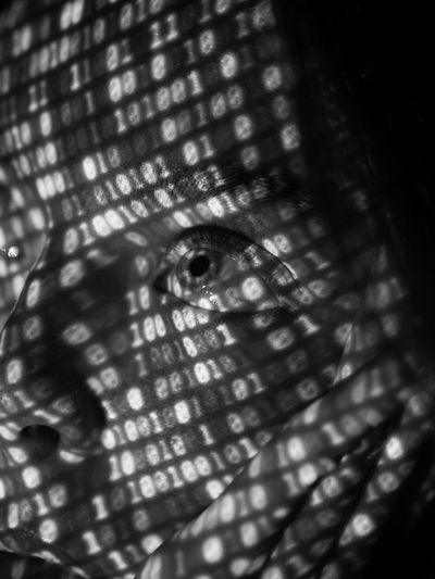 Close-up of a face with binary code projected onto it, creating a pattern of light and shadow across the skin and eye. Black and white.