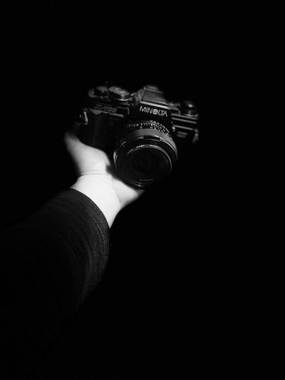Une main tenant un appareil photo Minolta vintage sur un fond noir, éclairée de manière spectaculaire pour mettre en valeur les détails de l'appareil photo.