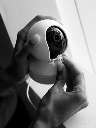Hands inserting a microSD card into a round security camera, positioned near a window. Black and white image.