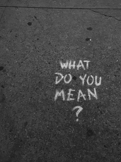 Image en noir et blanc d'un graffiti sur le trottoir indiquant « QU'EST-CE QUE TU VEUX DIRE ? » incitant à la réflexion ou à la méditation.