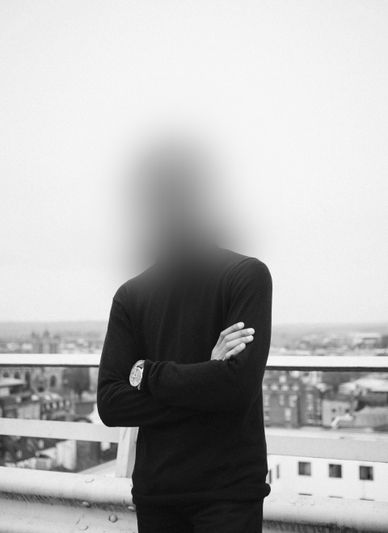 Une personne au visage flou se tient sur un toit, portant un pull sombre et une montre, avec un paysage urbain en arrière-plan. Image en noir et blanc.