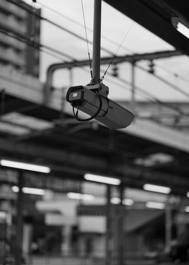 Image en noir et blanc d'une caméra de surveillance suspendue au plafond d'une gare, avec des éléments structurels flous en arrière-plan.