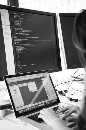 Persona escribiendo en una laptop con código en varios monitores de fondo, creando un ambiente de trabajo tecnológico enfocado. Imagen en blanco y negro.