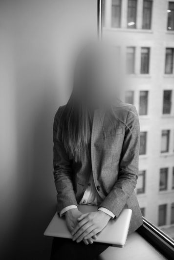 Personne en costume assise près d'une fenêtre avec un ordinateur portable, visage flou, et un bâtiment de la ville visible à l'extérieur. Image en noir et blanc.