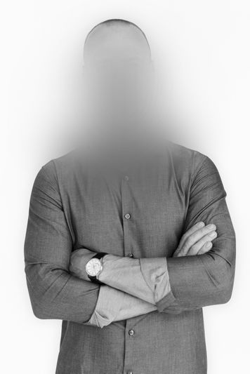 Man with blurred face, bald head, wearing a buttoned shirt and watch, standing with arms crossed against a plain background.