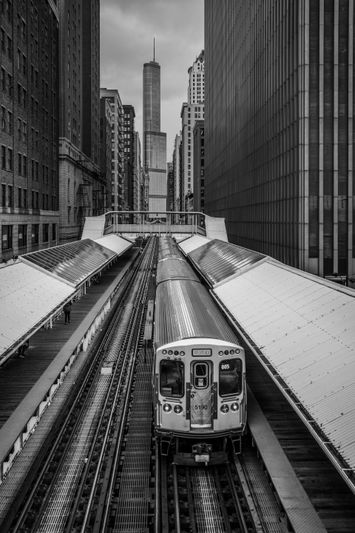 Image en noir et blanc d'un train à une station élevée en ville avec des gratte-ciel en arrière-plan.