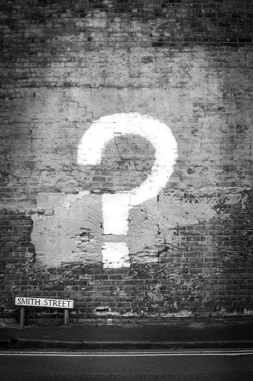 Un grand point d'interrogation blanc peint sur un mur de briques au-dessus d'un panneau « Smith Street ». L'image est en noir et blanc.