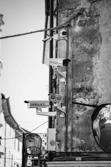 Plusieurs caméras de surveillance montées sur un mur patiné, avec un miroir convexe reflétant une rue étroite en noir et blanc.