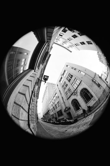 Fisheye lens view of an urban street, showing tall buildings and a cobblestone path, creating a curved and distorted perspective. Black and white.