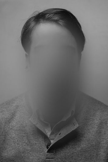 A grayscale portrait of a person with a blurred face, wearing a collared shirt and sweater, set against a plain background.