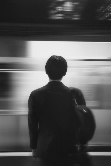 Une personne en costume avec un sac à bandoulière se tient sur un quai, face à un train flou et rapide dans un décor monochrome.