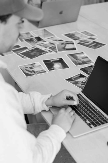 Persona con gorra usando una computadora portátil en una mesa, rodeada de fotografías en blanco y negro dispersas.