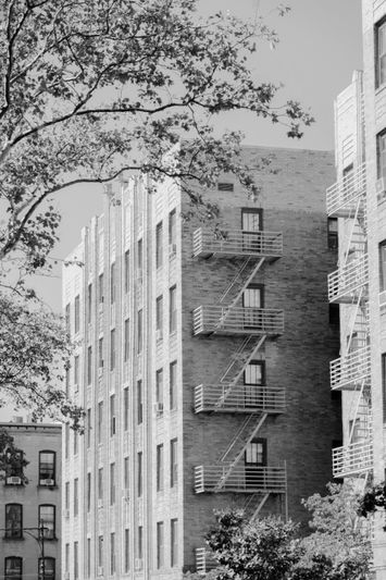 Photo en noir et blanc d'un immeuble en briques à plusieurs étages avec des escaliers de secours extérieurs, partiellement encadré par des arbres, sous un ciel dégagé.