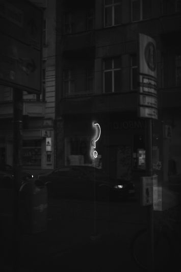 Neonowy znak zapytania świeci w ciemnym miejskim otoczeniu, otoczony słabo oświetlonymi budynkami i zaparkowanym samochodem na ulicy.