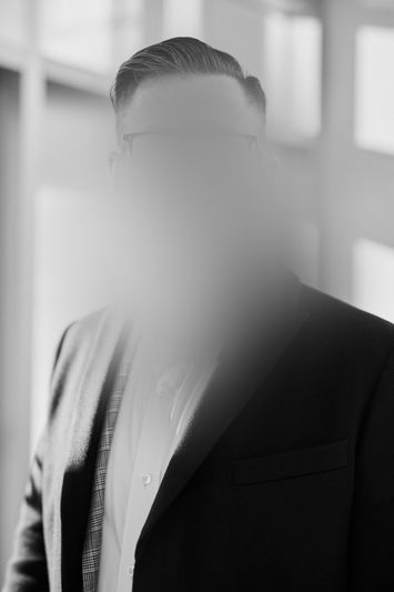 Homme en costume au visage obscurci, debout à l'intérieur près de grandes fenêtres, éclairé par la lumière naturelle. Photo en noir et blanc.