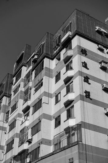 Image en noir et blanc d’un immeuble de plusieurs étages présentant des motifs de carreaux rayés, des balcons et des climatiseurs visibles, sous un ciel dégagé.