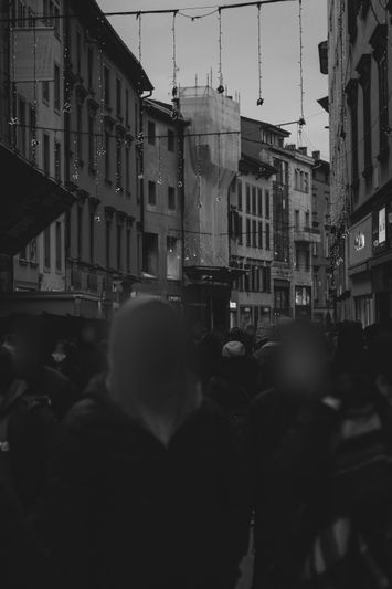 Rue animée d'une ville avec de vieux bâtiments, des lumières suspendues et des passants. La scène est en noir et blanc.