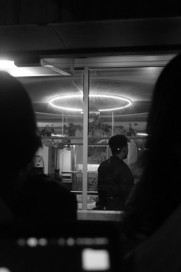 Una persona está de pie dentro de un diner, vista a través de una ventana por la noche. Un dispositivo de luz circular brillante cuelga del techo, creando un efecto de halo.