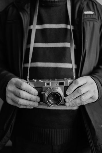 Person holding a vintage camera with both hands, wearing a striped sweater and jacket. Black and white image.