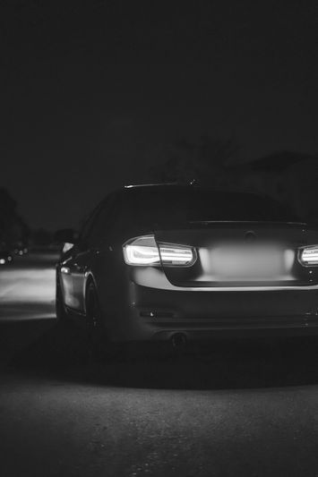 Black and white image of a car with illuminated taillights, parked on a dimly lit street at night.