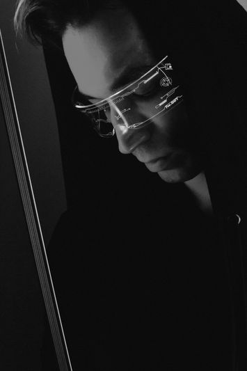 Person in a dark hoodie wearing futuristic, illuminated glasses, looking down in a dimly lit environment.